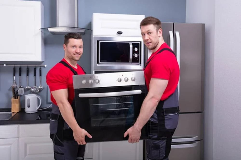Two professional movers providing appliance removal services in San Antonio, TX by carrying a built-in oven from a residential kitchen.